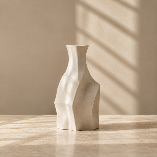 Ivory sculptural vase with angular facets and a narrow neck on a sunlit tabletop.