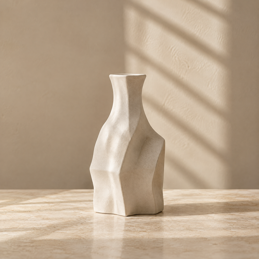 Ivory sculptural vase with angular facets and a narrow neck on a sunlit tabletop.